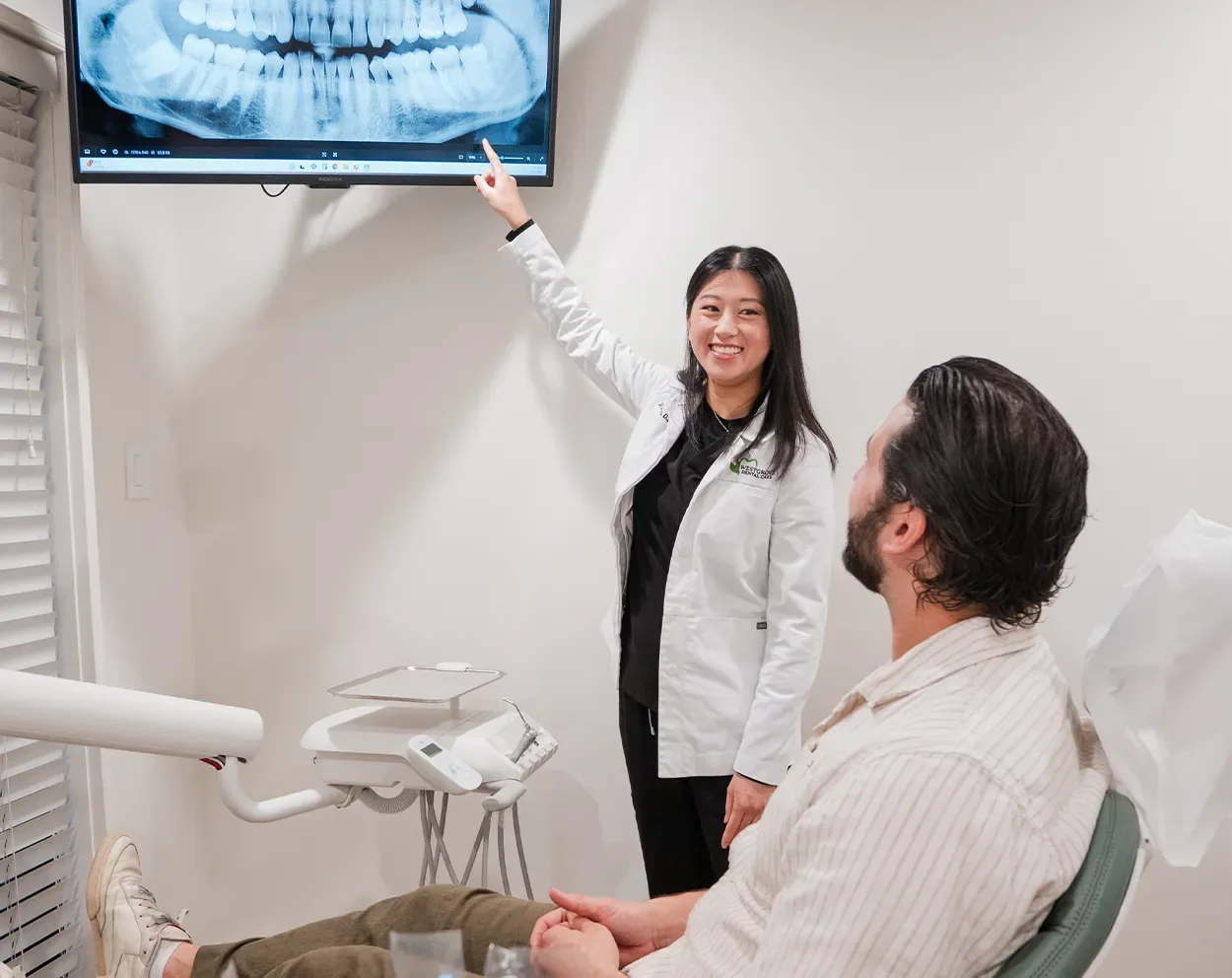 A dentist pointing to a digital X-ray on the wall monitor while a patient looks on